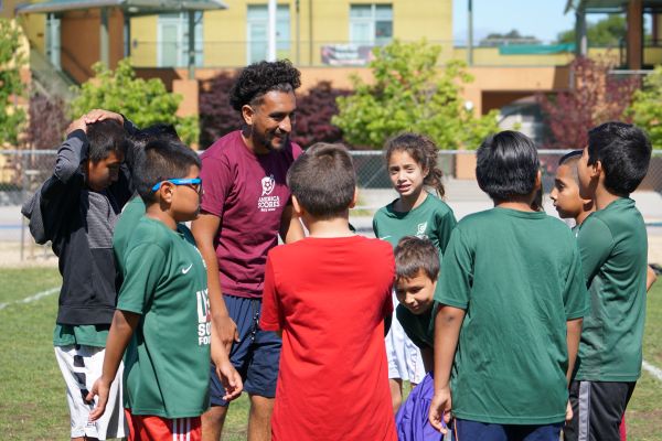 A male coach in a maroon U.S. Soccer Foundation shirt smiles as he gathers a diverse group of young children in green soccer jerseys in a huddle on a grassy field. They appear engaged and happy, with a school building and trees in the background.